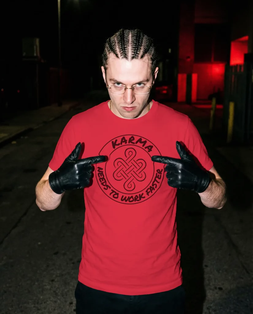 Homme en tee-shirt rouge pointant du doigt un logo circulaire "Karma needs to work faster" orné d'un nœud celtique, gants en cuir noir, ambiance urbaine nocturne