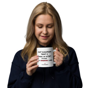 Tasse blanche avec inscription noire « most Europeans treat a Made in U.S.A. sticker as a » et mot WARNING en rouge.